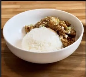 White rice with flavorful stir-fried chicken and vegetables in a white bowl on a wooden table.